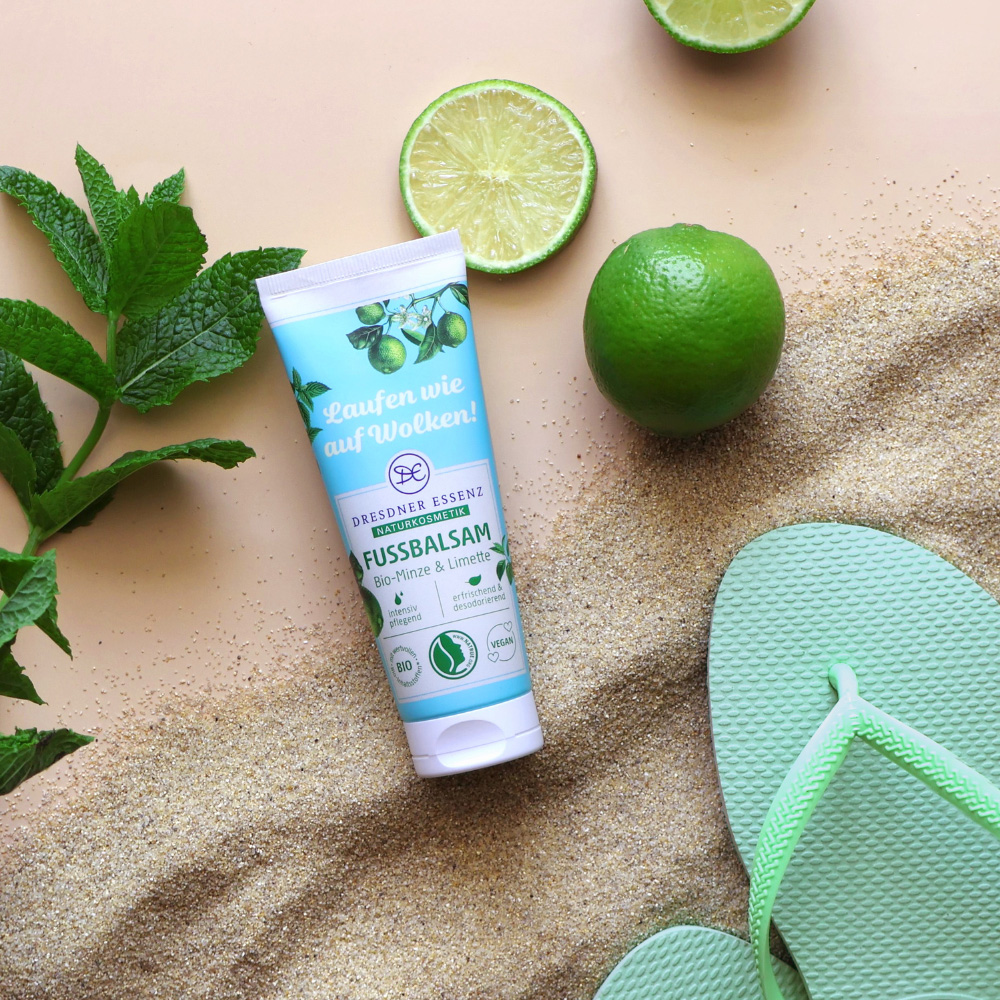 Der Fussbalsam liegt im sand und ist drum herum dekoriert mit Limetten, Minzblaettern und Flip-Flops.