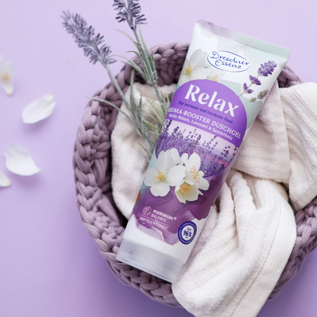 Das Aroma-Booster Duschgel relax liegt in einem violetten Korb mit einem weissen Handtuch, Lavendel & weissen Blueten.