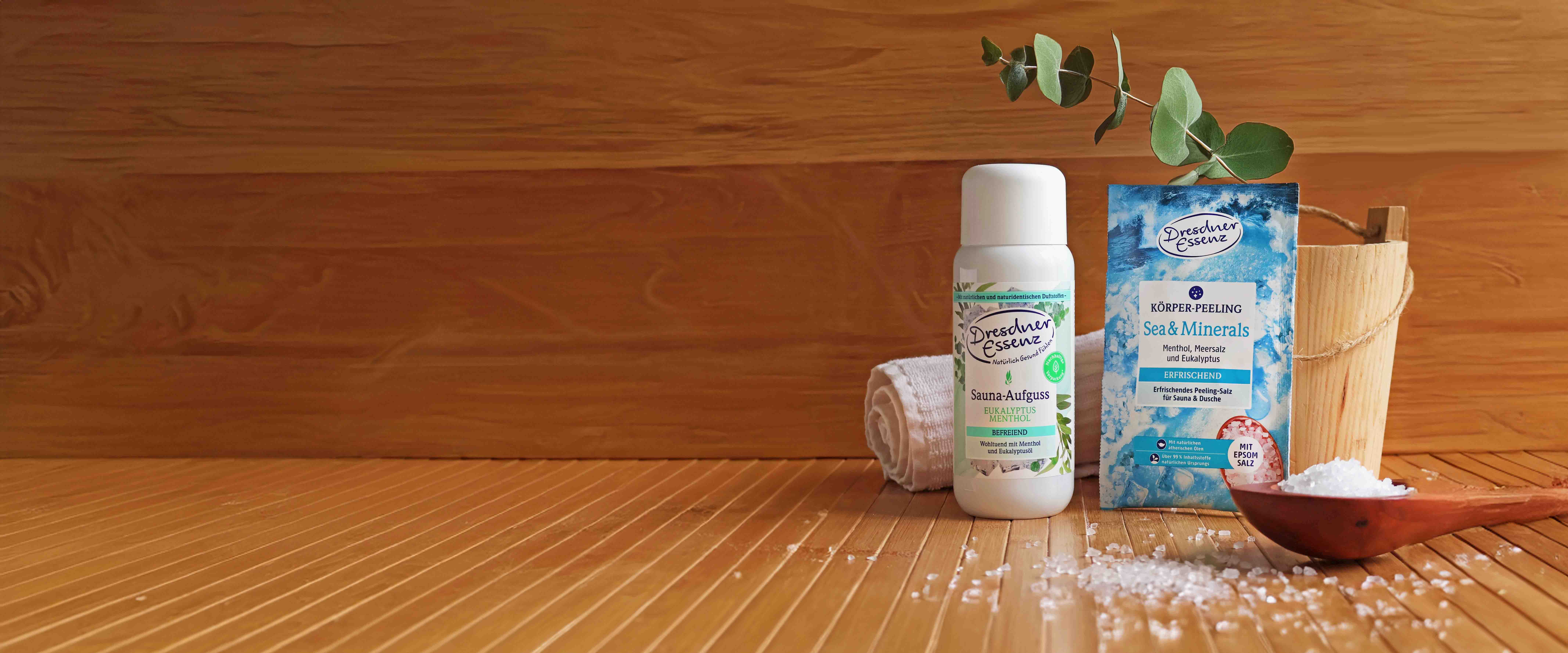 Sauna Aufguss Eukalyptus Menthol und Koerper peeling Sea & Minerals stehen in einer Sauna neben einem Sauna-Eimer, einem Holzloeffel mit Badesalz und einem Handtuch.