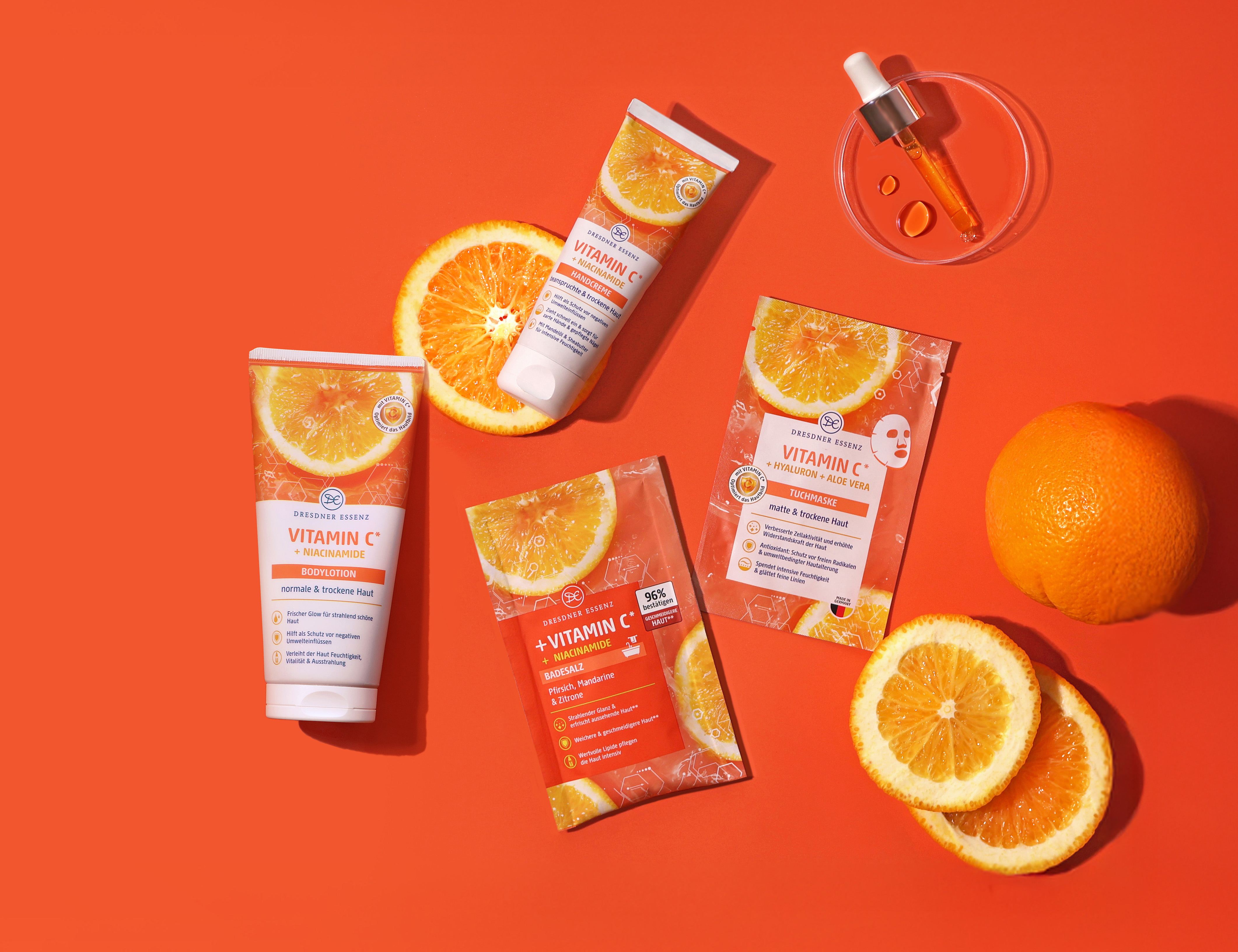 Die Produkte Badesalz, Tuchmaske, Handcreme und Bodylotion aus der Vitamin C Serie liegen nebeneinander und drum herum liegen dekorativ Orangen und Orangenscheiben und eine Pipette.