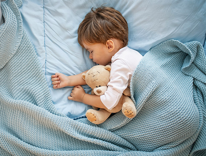 ein kleines Kind hat einen Teddybaeren im Arm und schlaeft in einer Decke eingekuschelt