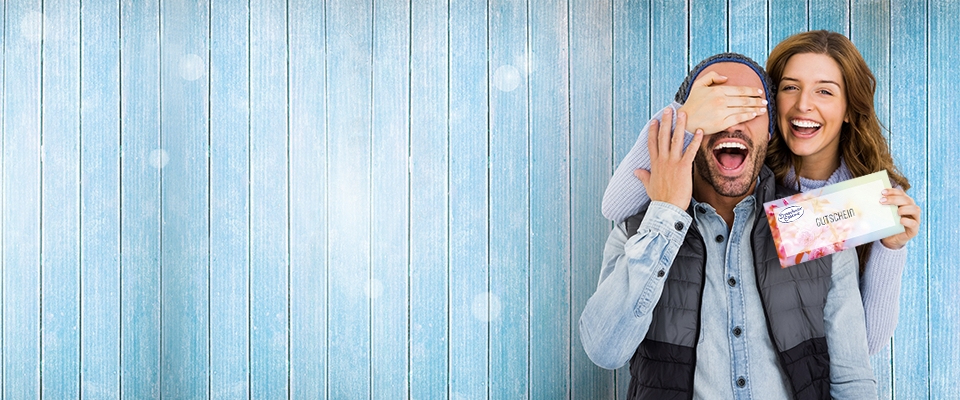 Ein Mann und eine Frau stehen vor einer blauen Wand. Die Frau verdeckt die Augen des Mannes und zeigt eine Gutschein-Karte.