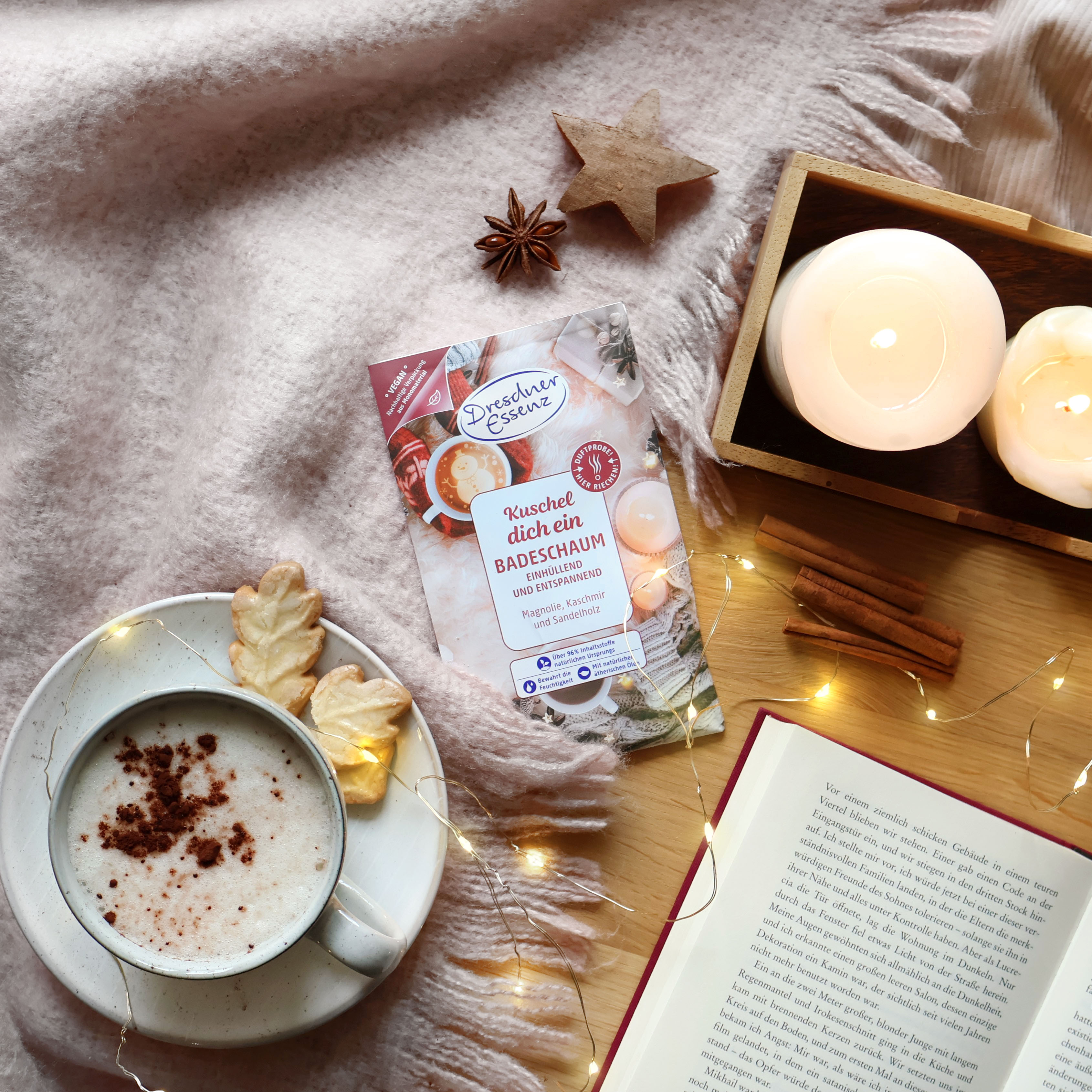 Badeschaum Kuschel dich ein liegt auf einer Kuscheldecke und drum herum steht eine Tasse Kakao, Kerzen, ein Buch, eine Lichterkette und verschiedene Gewuerze