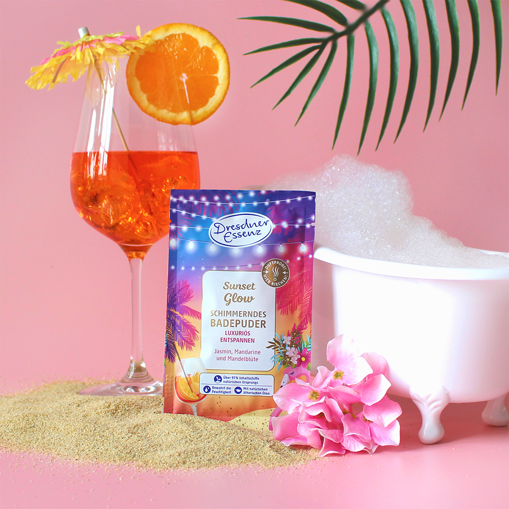 Badepuder Sunset Glow steht im Sand neben einer kleinen Badewanne und einem frischen Cocktail.