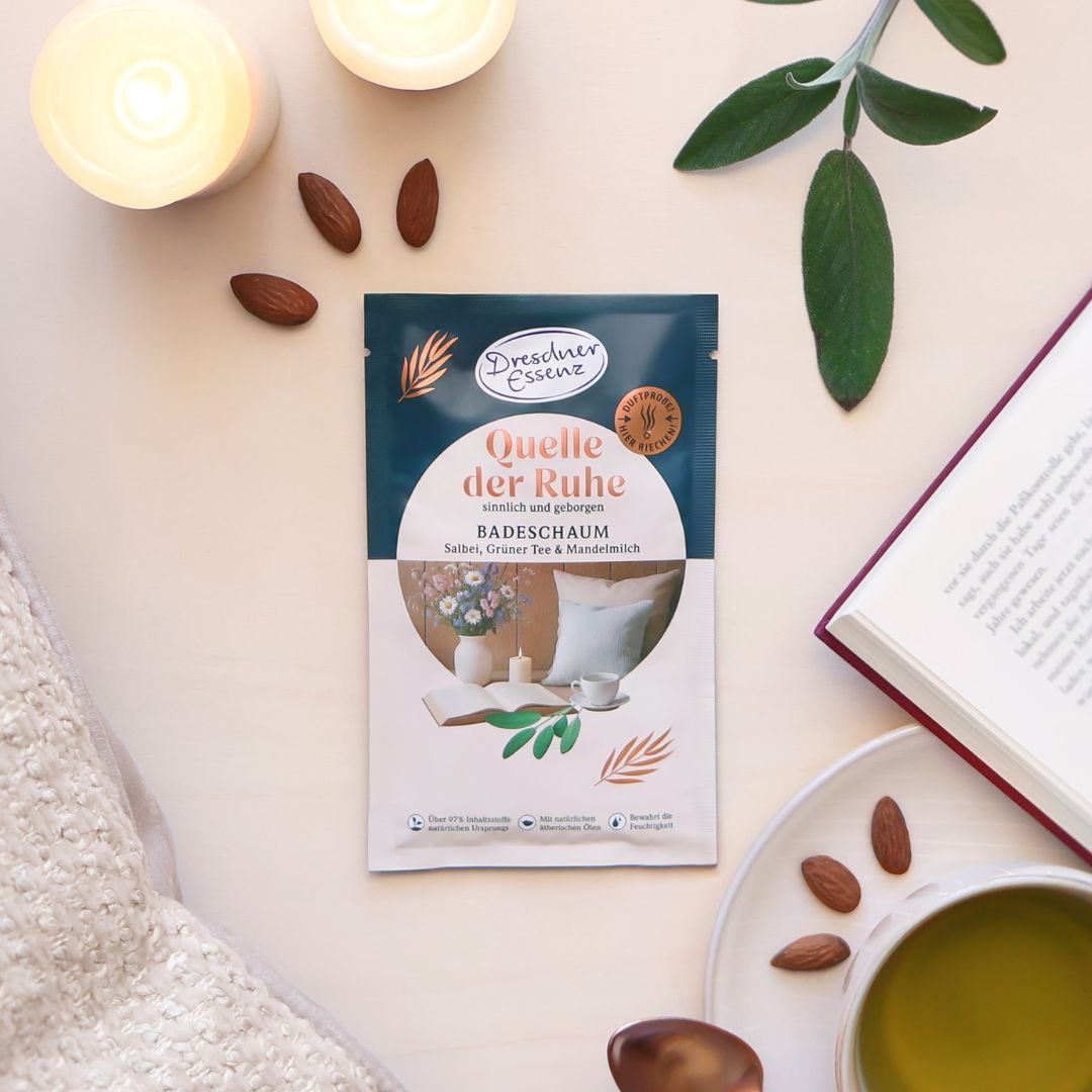 Der Badeschaum Quelle der Ruhe ist drum herum mit Mandeln, Salbei, einem Buch, Kerzen und einer Tasse Tee dekoriert.