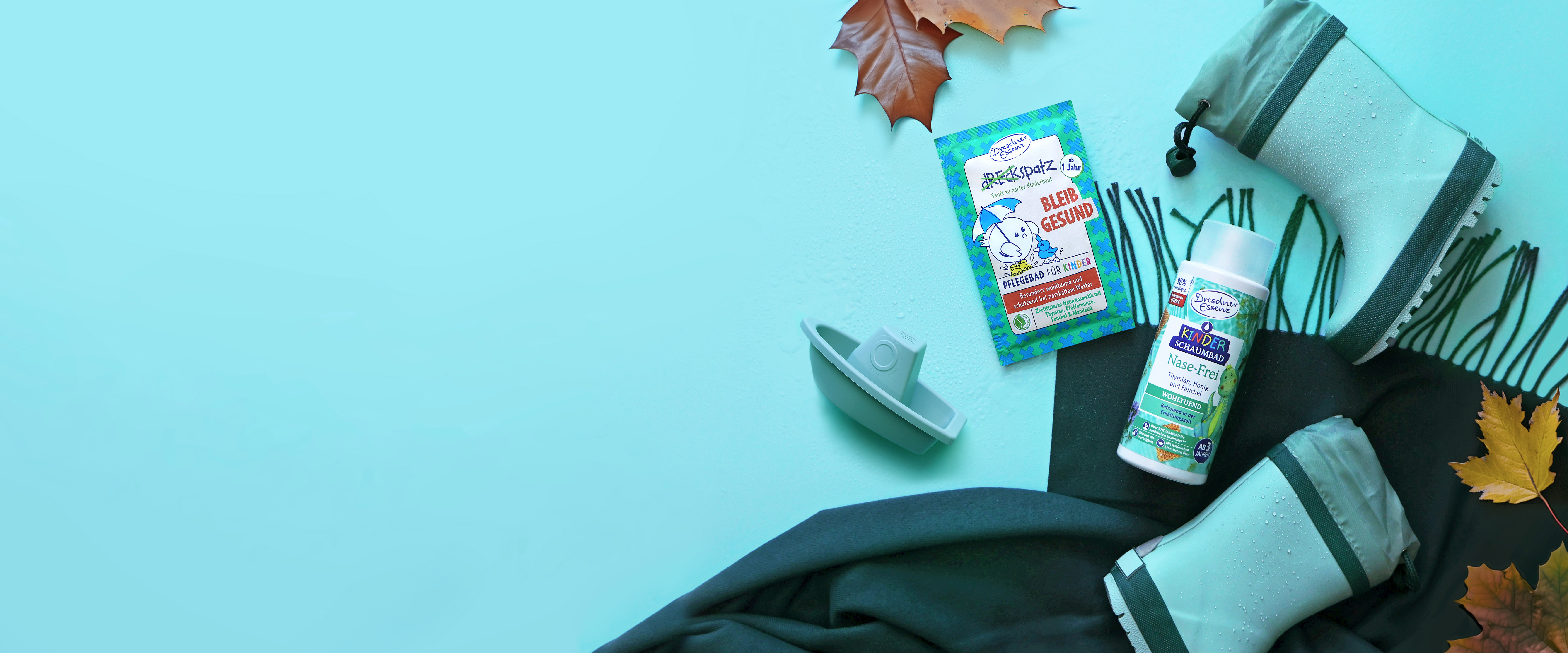 Das Pflegebad Bleib gesund und das Kinder-Schaumbad Nase Frei liegen nebeneinander auf einem dicken Schal und sind drum herum dekoriert mit Gummistiefeln, einem kleinen Spielzeug-Boot und Herbstblaettern.
