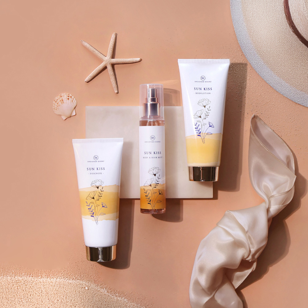 Sun Kiss Produkte liegen nebeneinander und sind dekoriert mit Sand, Muscheln und Seesternen sowie eleganten Stoffen