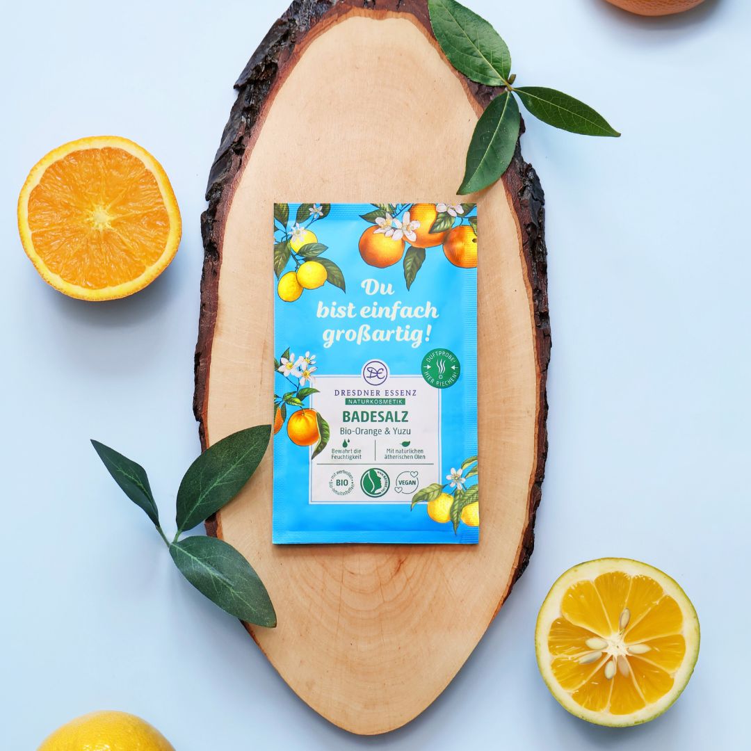 Das Badesalz Bio-Orange und Yuzu liegt auf einem Stueck Holz und ist mit Orangen und frischen Blaettern dekoriert.