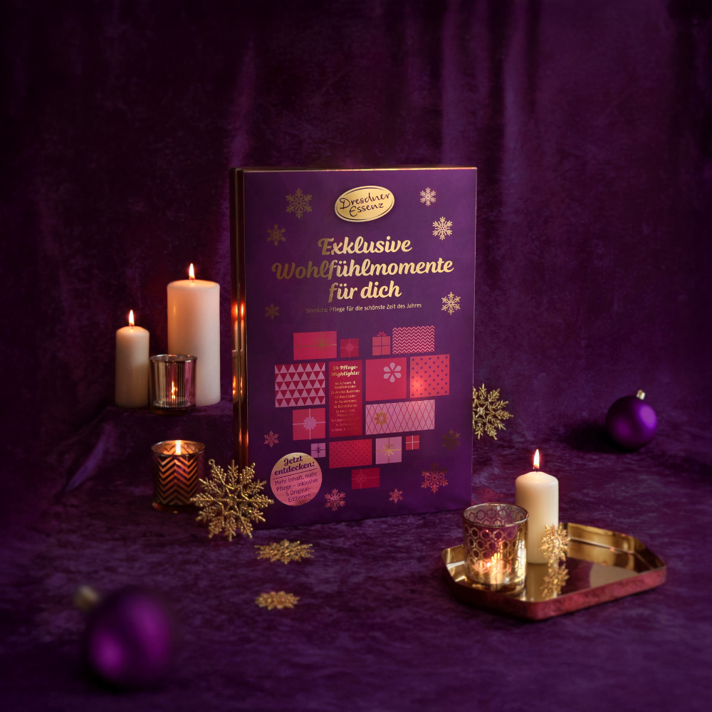 Adventskalender steht im Fokus und ist drum herum dekoriert mit goldenen Elementen, Kerzen, Christbaumkugeln und violettem Stoff