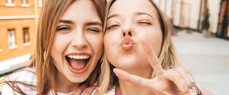 Zwei Teenager Maedchen stehen nebeneinander und lachen. Ein Maedchen zeigt ein Peace-Zeichen.