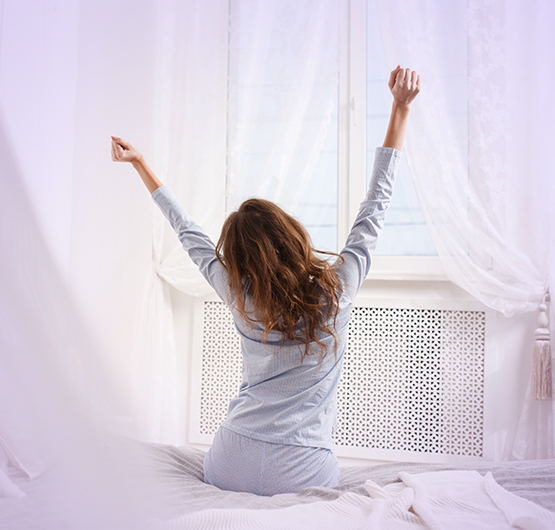 Eine Frau sitzt am Bettenrand und streckt sich und schaut dabei aus dem Fenster.