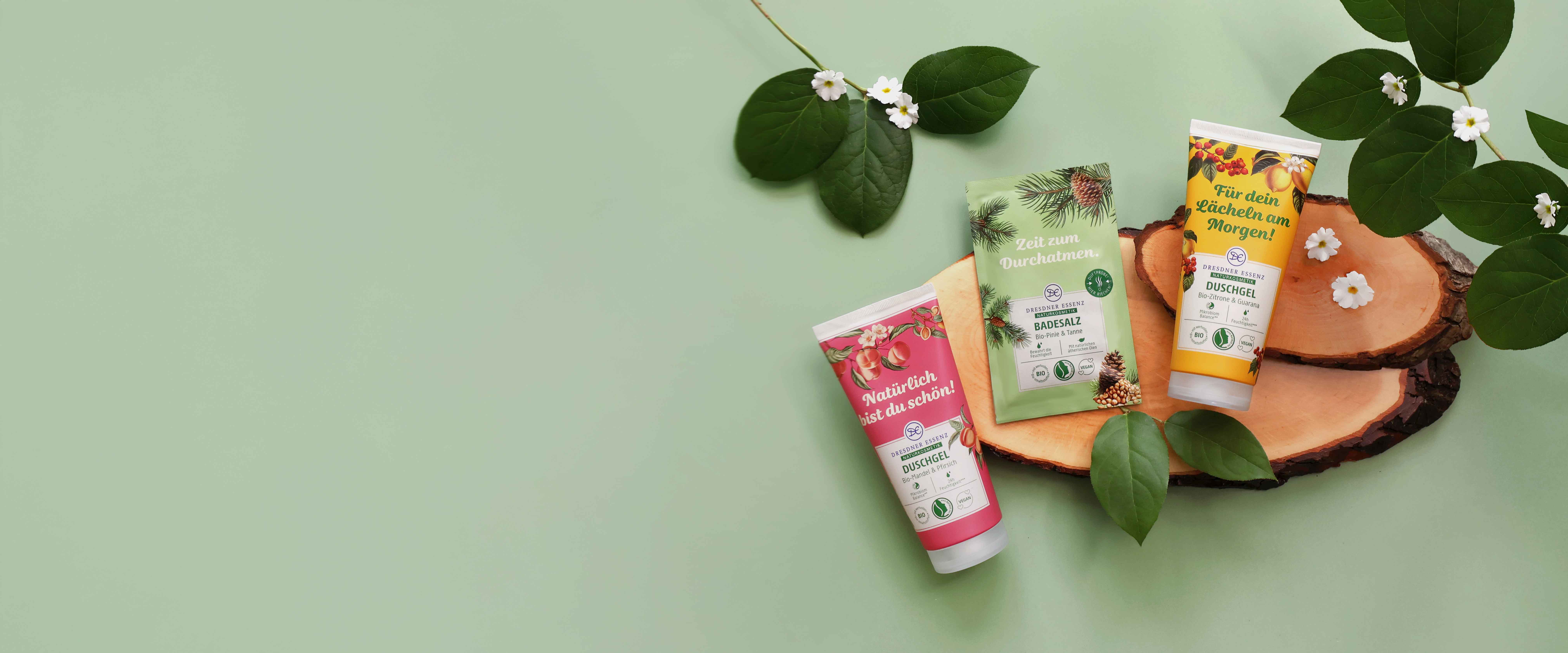 Die zwei Duschgele "Natürlich bist du schön" und "Für dein Lächeln am Morgen" und das Badesalz "Zeit zum Durchatmen" der Naturkosmetik-Linie liegen nebeneinander. Um sie herum Deko-Elemente wie Holz, Blüten und Blätter