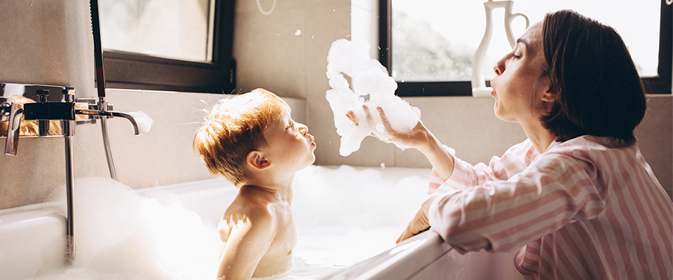 Ein Kind sitzt in der Badewanne und eine Frau am Rand der Wanne. Die Frau hat Schaum in der Hand und beide pusten den Schaum in die Luft.