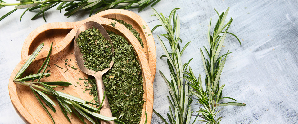 Nahaufnahme verschiedener Rosmarin-Zweige und getrocknetem Rosmarin, der in einer Holzschale und einem Holzloeffel liegt