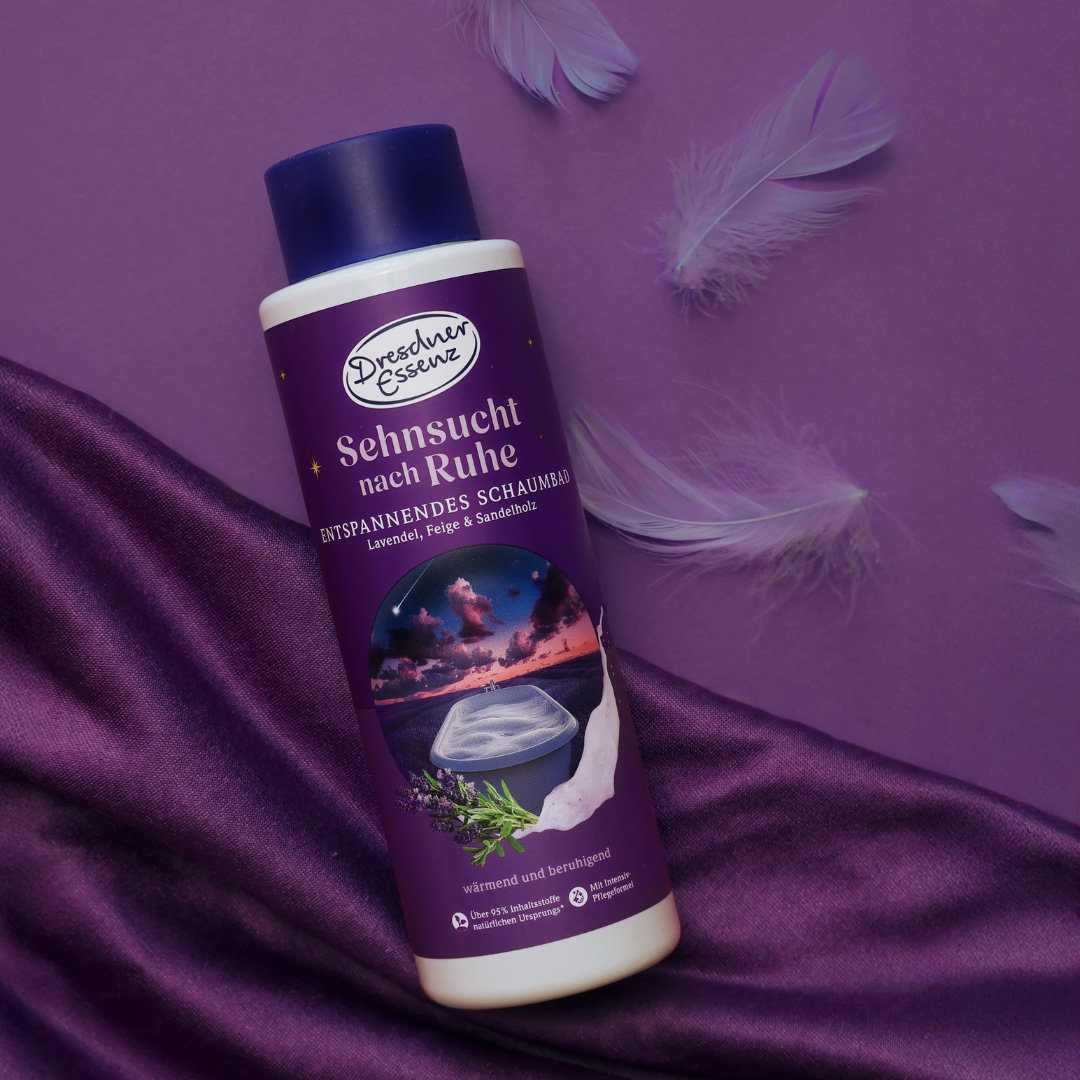 Das Schaumbad Sehnsucht nach Ruhe liegt auf einem dunkel-violetten Tuch und ist dekoriert mit violetten Federn.
