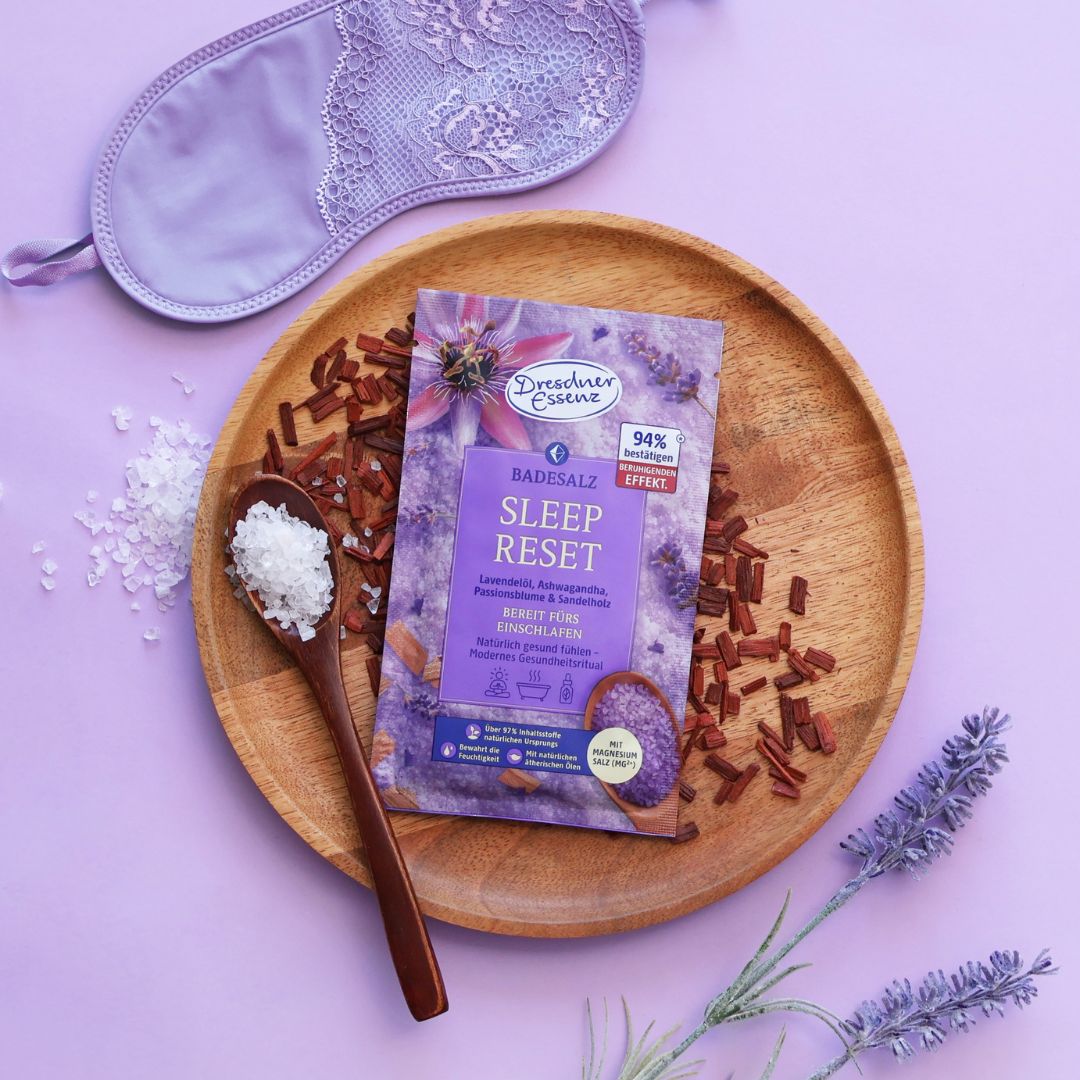 Das Badesalz Sleep Reset liegt auf einem Holzteller und ist mit einer Schlafmaske, Lavendel und Sandelholz dekoriert.