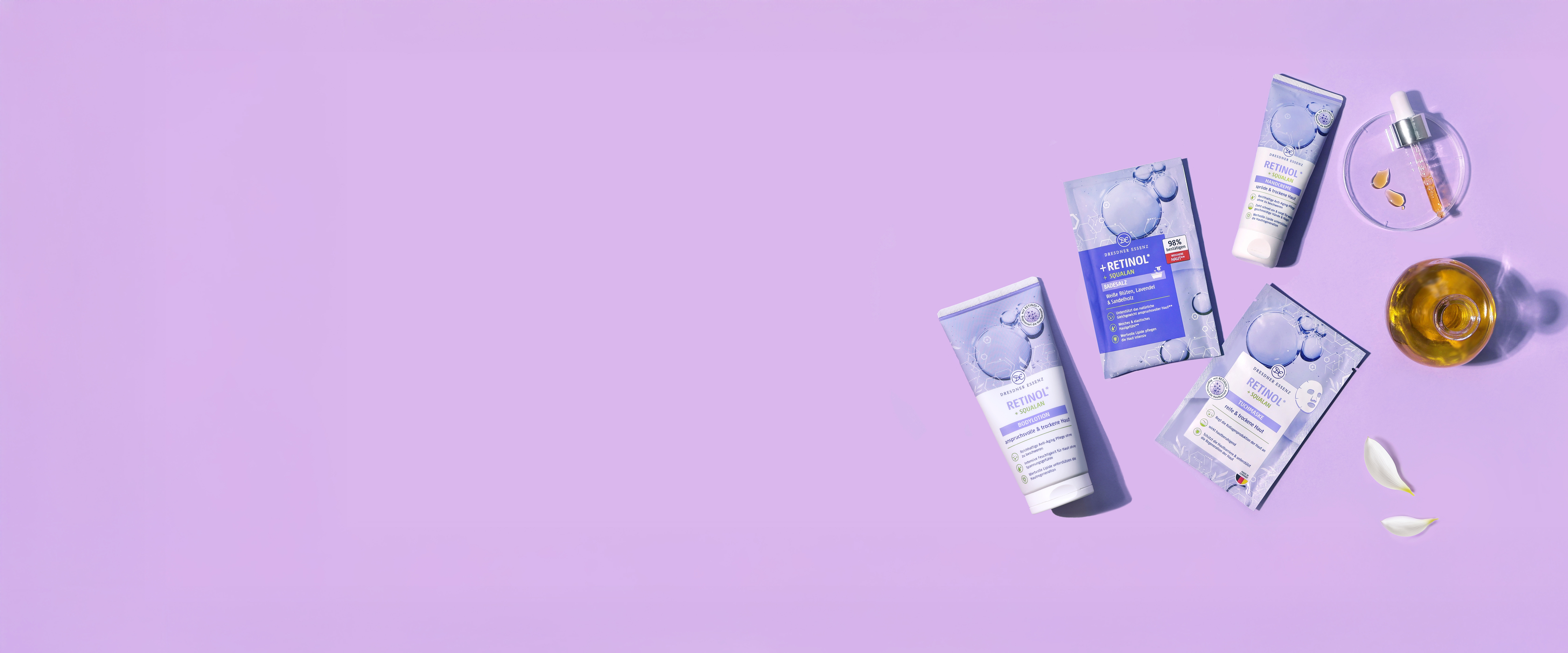 Die Produkte Badesalz, Tuchmaske, Handcreme und Bodylotion aus der Retinol Serie liegen nebeneinander und drum herum liegen dekorativ Blueten und eine Pipette.