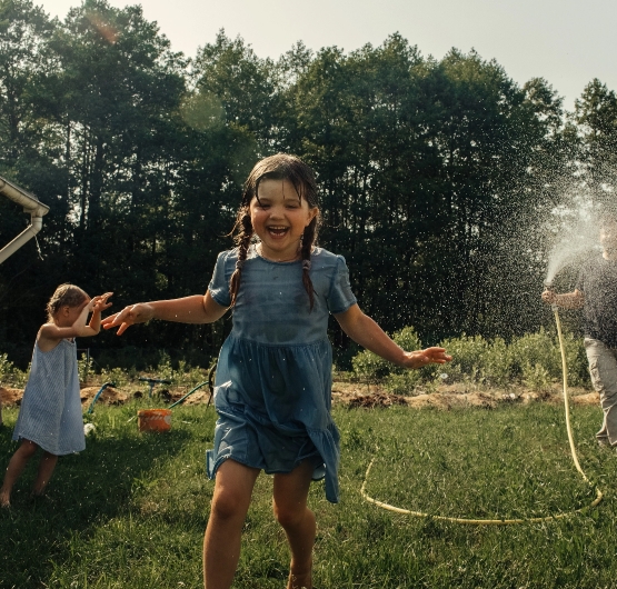 Zwei Kinder spielen im Sommer draussen im Garten. Ein Erwachsener spritzt sie mit einem Gartenschlauch nass.