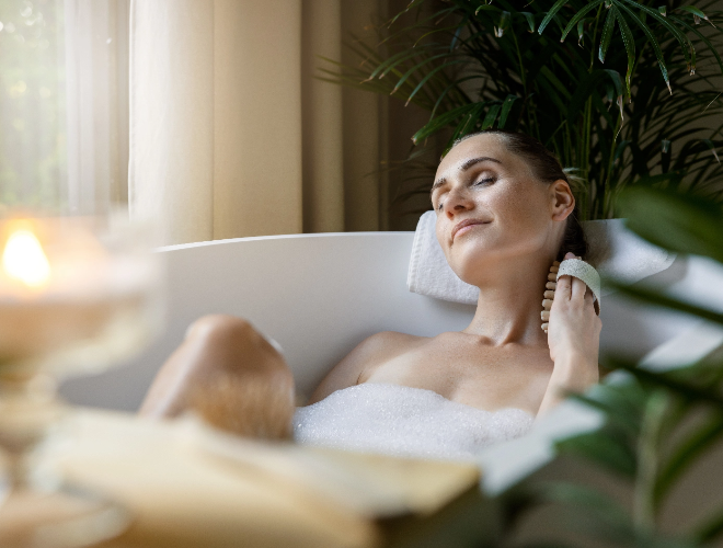 Eine Frau liegt mit geschlossenen Augen in der Badewanne und streicht sich mit einer Buerste ueber den Hals und entspannt sich.