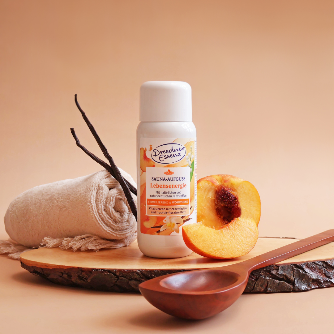 Der Sauna Aufguss Lebensenergie steht auf einem Holzbrett und ist ringsherum dekoriert mit Vanillestangen, Pfirsich, einem Handtuch und einer Sauna-Kelle.