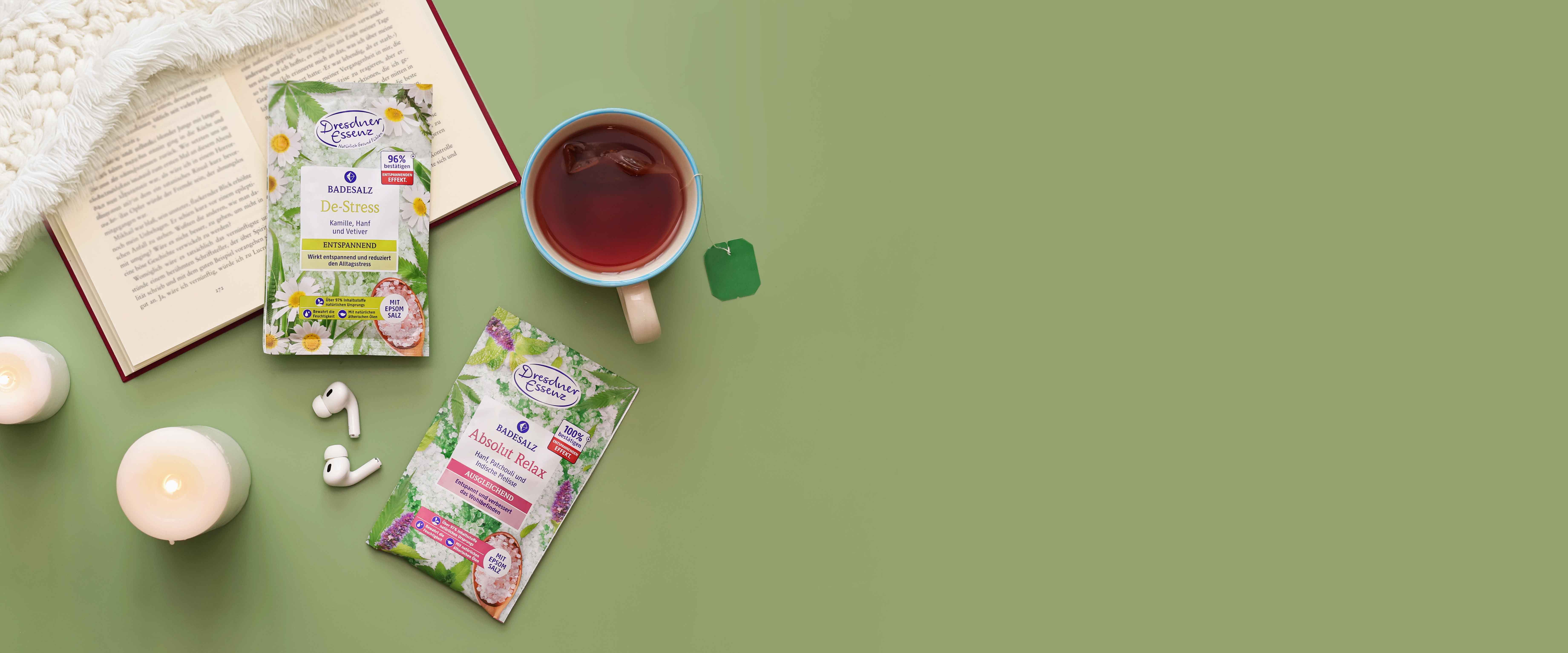 Die Badesalze Absolut Relax und De-Stress liegen nebeneinander und sind ringsherum dekoriert mit einer Kuscheldecke, einem Buch, Kerzen und einer Tasse Tee.