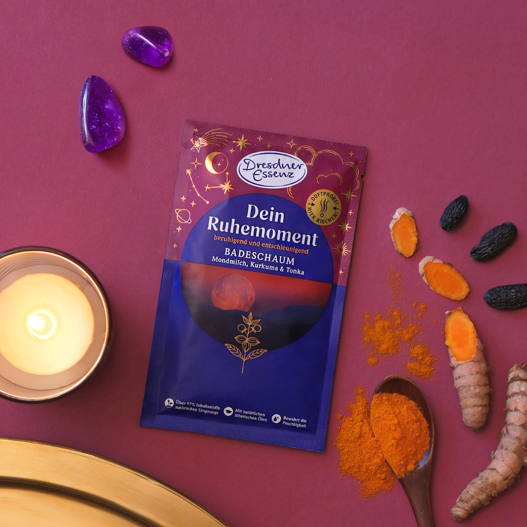 Der Badeschaum Dein Ruhemoment liegt im Fokus und ist drum herum dekoriert mit Kurkuma, Tonkabohnen, Kerzen, violetten Steinen und Gold-Elementen.