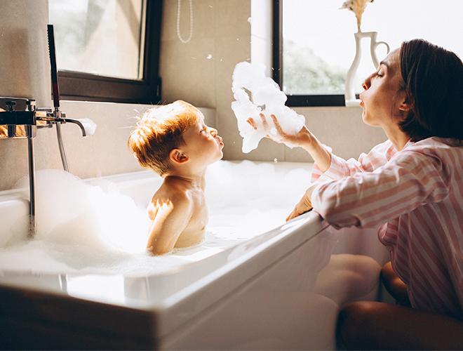 Ein Kind badet in einer schaeumenden Badewanne. Am Rand der Wanne sitzt die Mutter und beide pusten den Schaum in die Luft.