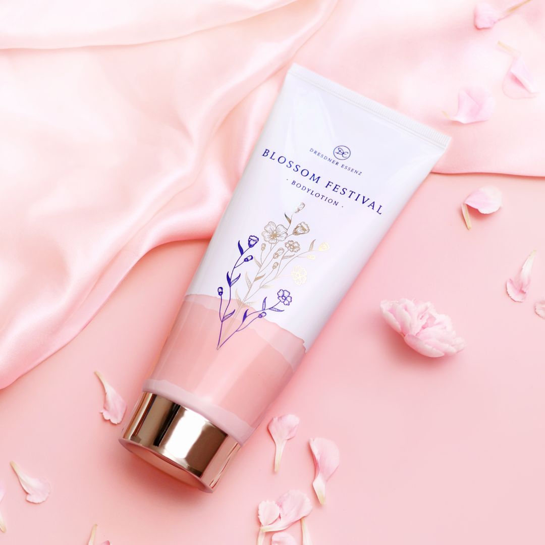 Die Bodylotion liegt vor einem Hintergrund in zart-rosa und ist dekoriert mit rosafarbenen Blueten und elegantem Satin-Stoff.