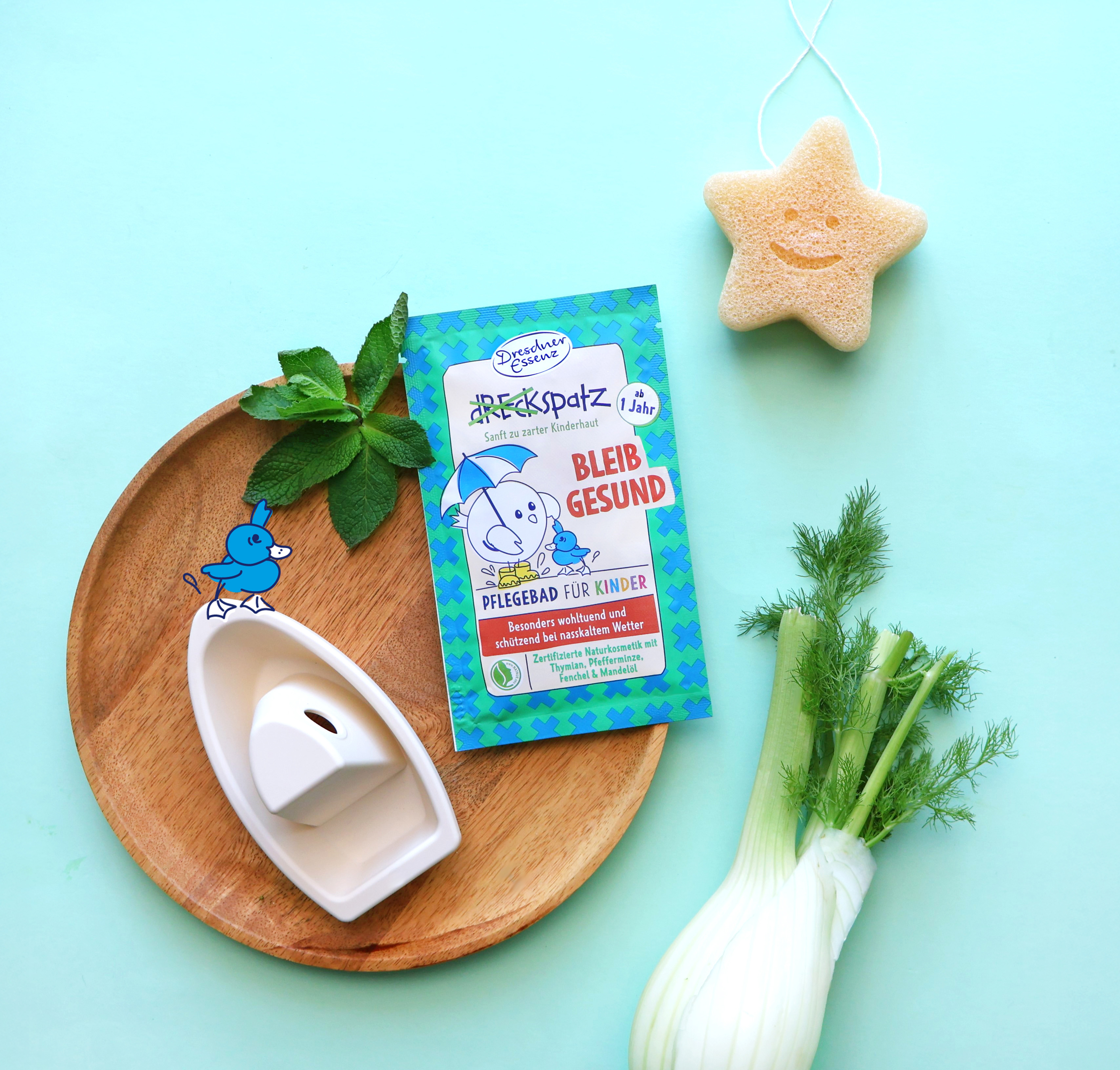 Pflegebad Bleib gesund liegt neben einem kleinen Spielzeugboot und Minze auf einem kleinen Holztablett. Neben dem Tablett liegt ein grosser Fenchel und ein kleiner Schwamm in Sternen-Form.