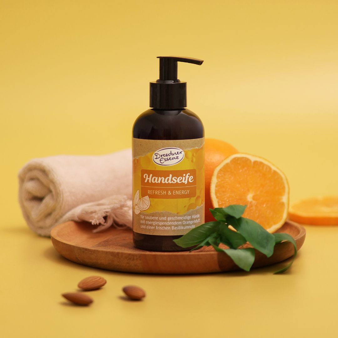 Die Handseife Refresh and Energy steht auf einem Holztablett, umgeben von Orangen, Basilikum, Mandeln und einem Handtuch.