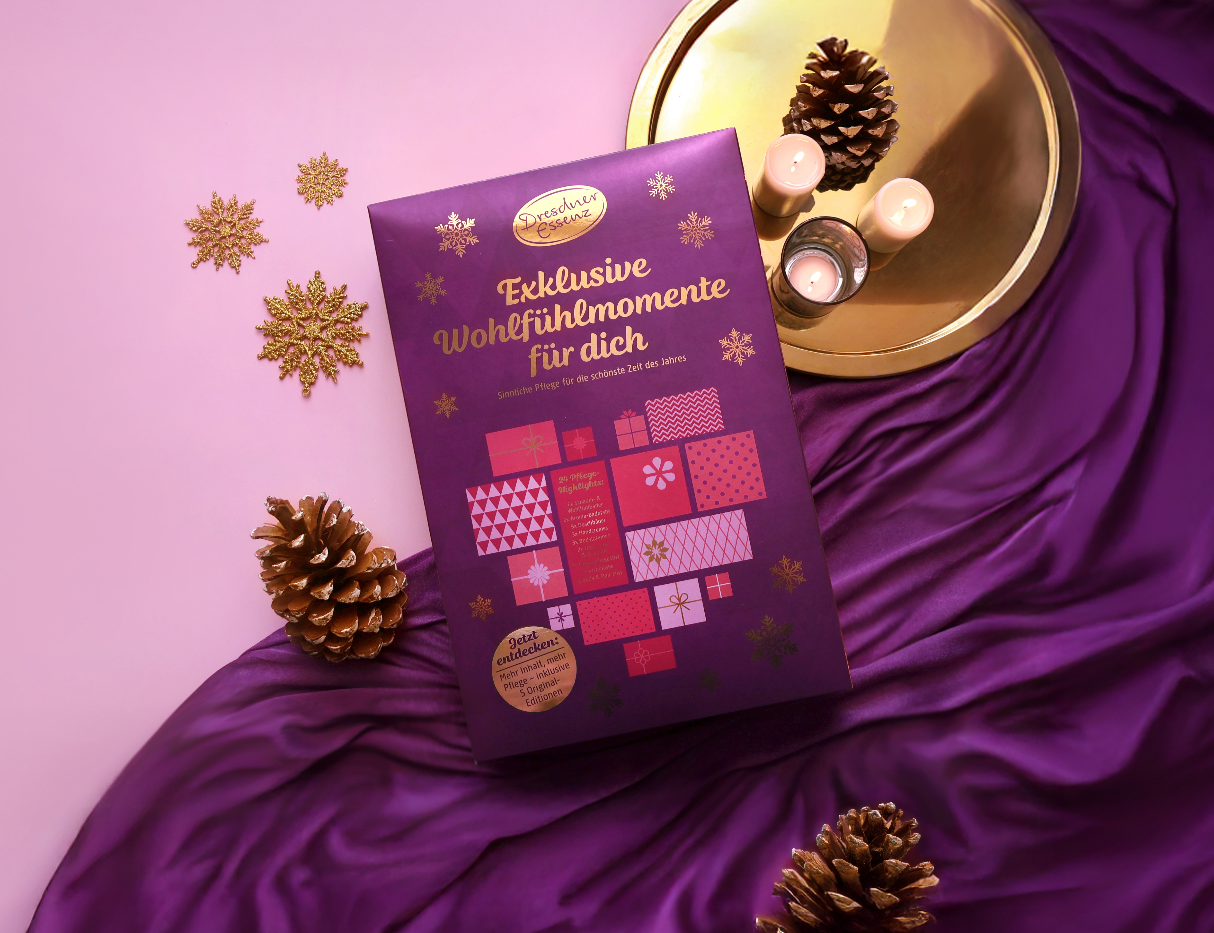 Der Adventskalender 2025 liegt auf einem violetten Samt-Tuch und ist ringsherum dekoriert mit Kerzen und goldenen Schneeflocken.