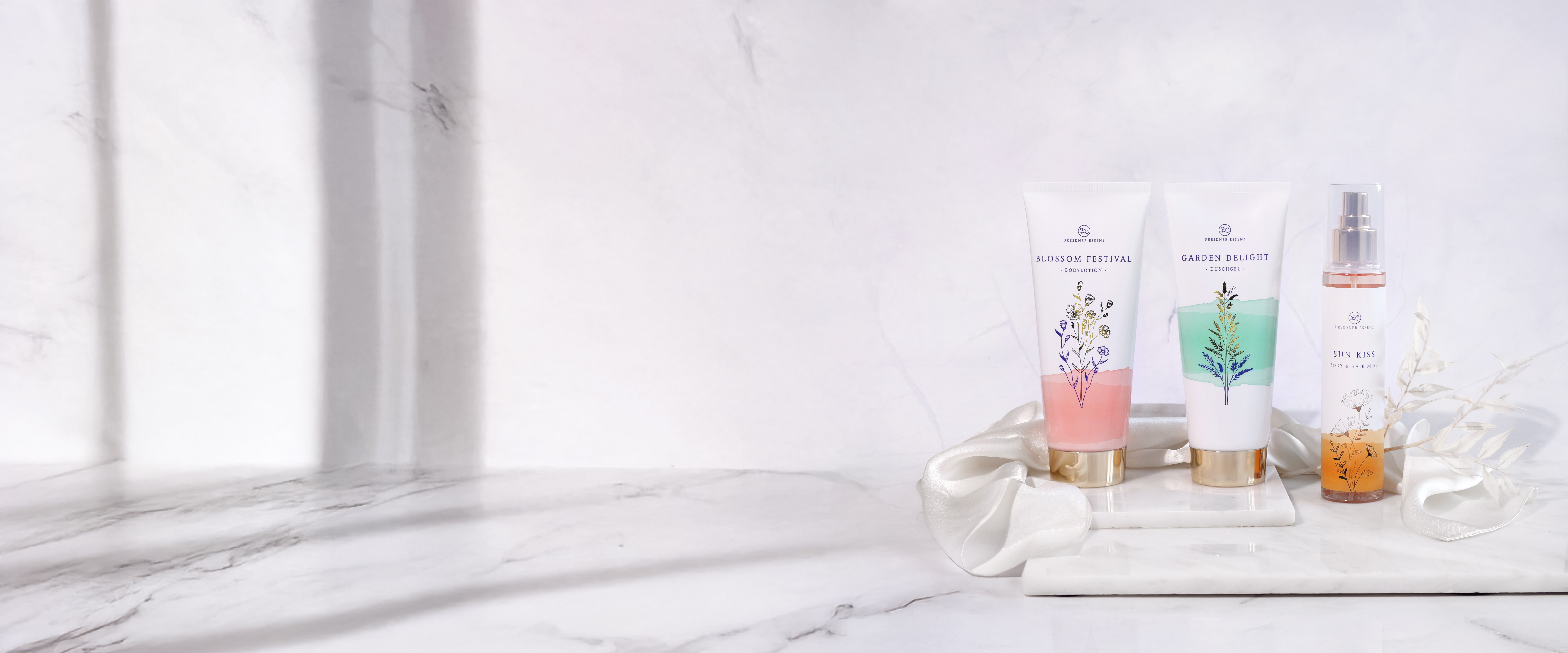 Die Bodylotion Blossom Festival, das Duschgel Garden Delight und das Body & Hair Mist Sun Kiss stehen nebeneinander vor einem edlen Marmorhintergrund und sind dekoriert mit weissen Blueten und edlem Satin-Stoff.