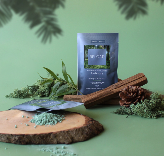 Das Badesalz RELOAD steht zwischen verschiedenen Blaettern, Tannenzapfen und Hoelzern. Im Vordergrund liegt ein weiteres Sachet des Produktes und das Produkt ist auf einem Holztablett verstreut.