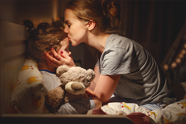 Ein Kind liegt eingekuschelt im Bett und die Mutter gibt ihm einen Gute-Nacht-Kuss auf die Stirn.