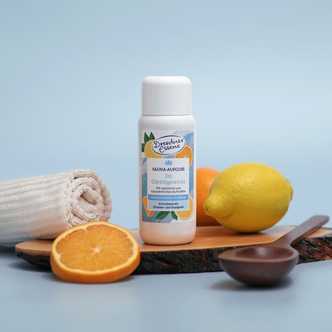Der Sauna Aufguss Im Gleichgewicht steht auf einem Holz-Stueck, umgeben von Orangen und Zitronen, einer Sauna-Kelle und einem Handtuch.