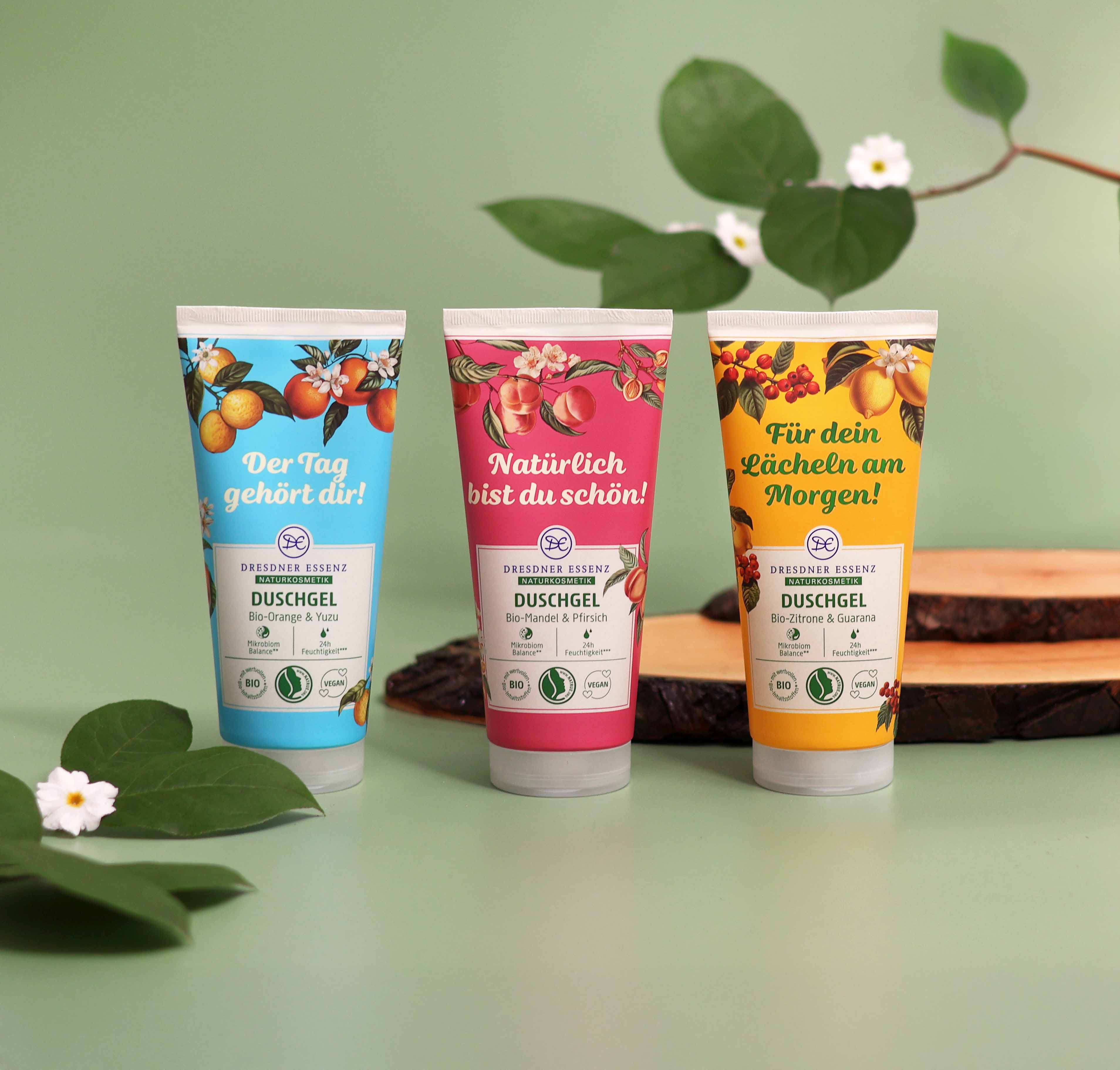 Die drei Duschgele "Der Tag gehört dir", "Natürlich bist du schön" und "Für dein Lächeln am Morgen" der Naturkosmetik-Linie stehen nebeneinander. Um sie herum Deko-Elemente wie Holz, Blüten und Blätter