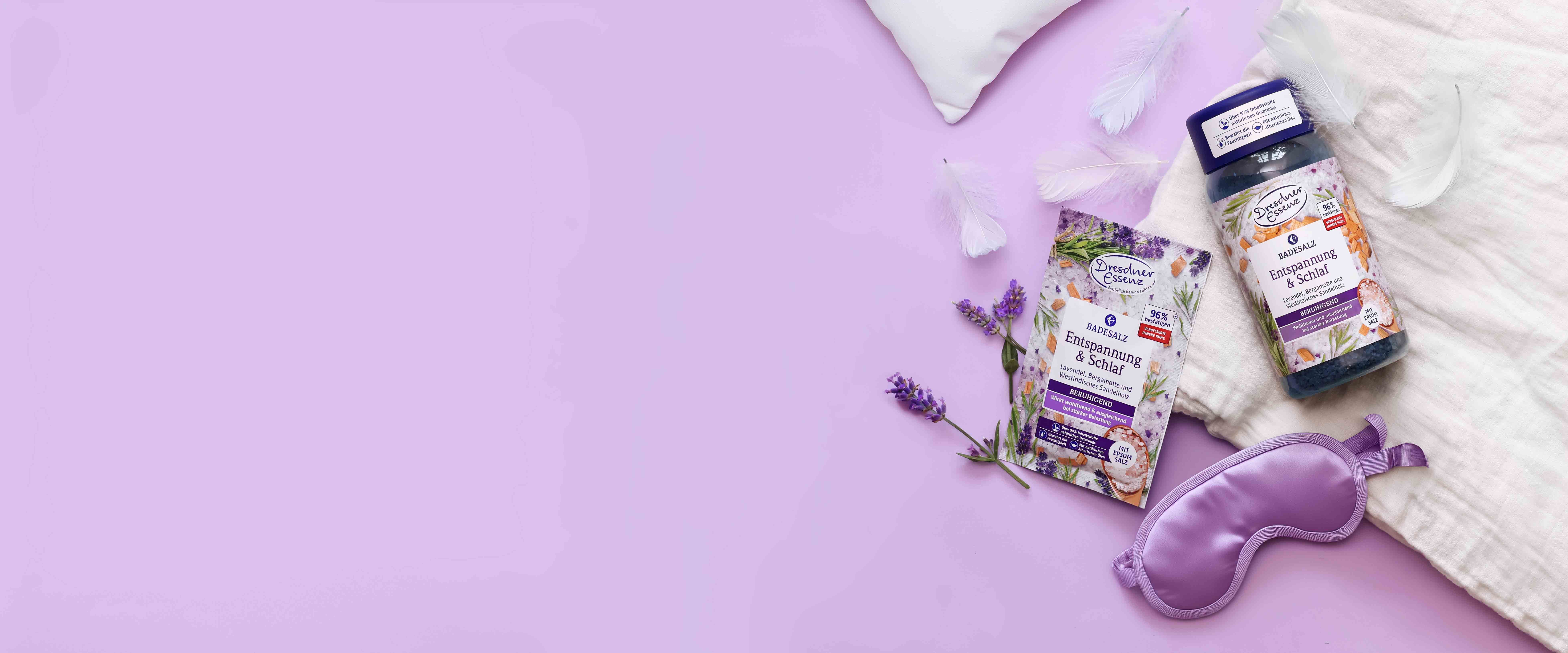 Das Badesalz Entspannung & Schlaf in der 60g Variante und in der 600g Dose liegen auf einem weichen Untergrund und sind rings herum dekoriert mit einer violetten Schlafmaske, Federn und Lavendel.